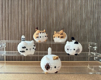 May include: Five handmade crochet cat figurines in white, brown, and black. The cat figurines are displayed on a clear acrylic shelf. The cats have small tails and are in a sitting position. The background is a brown, textured surface.
