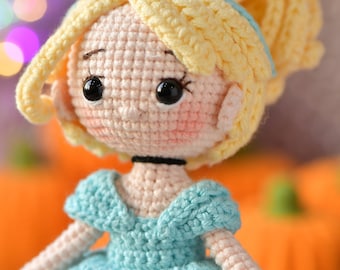 Könnte beinhalten: Gehäkelte Puppe einer Prinzessin mit blonden Haaren und einem blauen Kleid. Die Puppe hat einen hellen Hautton, rosige Wangen und schwarze Augen. Das Haar ist zu einem Dutt frisiert. Das Kleid hat eine weiße Bordüre. Der Hintergrund ist verschwommen mit Orange und Grün.