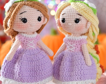 May include: Two handmade crochet dolls, each with a unique hairstyle and dress. One doll has brown hair, the other blonde. Both wear lavender dresses with pink bodices and white accents. The dolls are displayed on wooden stands.