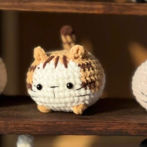 K&ouml;nnte beinhalten: Eine handgefertigte, geh&auml;kelte Katzenfigur in Wei&szlig;-, Beige- und Braunt&ouml;nen. Die runde Katze hat schwarze Augen und einen aufgestickten Mund. Die Katze sitzt auf einem Holzregal. Weitere &auml;hnliche Katzenfiguren sind im Hintergrund zu sehen.