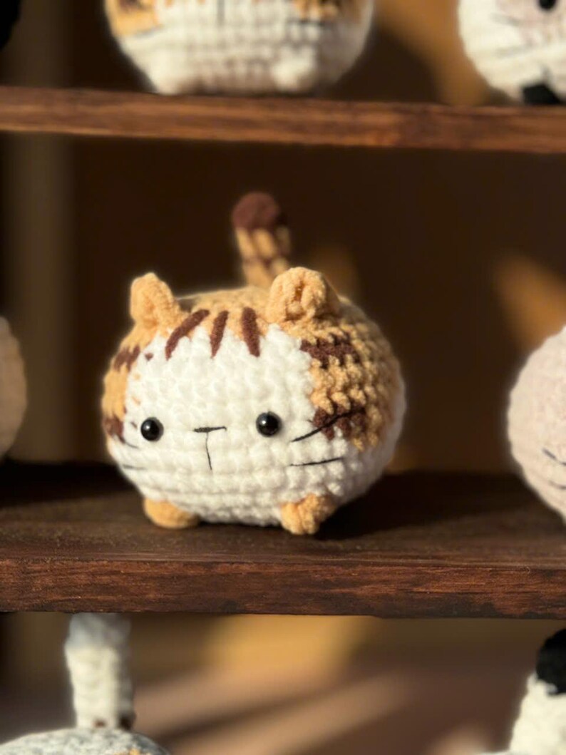 K&ouml;nnte beinhalten: Eine handgefertigte, geh&auml;kelte Katzenfigur in Wei&szlig;-, Beige- und Braunt&ouml;nen. Die runde Katze hat schwarze Augen und einen aufgestickten Mund. Die Katze sitzt auf einem Holzregal. Weitere &auml;hnliche Katzenfiguren sind im Hintergrund zu sehen.