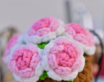 May include: A handcrafted crochet basket filled with pink and white flowers. The flowers have a detailed petal design, and the basket is a warm brown color. The text "Sunny Princess Crochet" is visible on the basket.