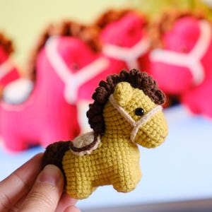 Puede incluir: Un juguete de caballo hecho a mano, tejido a ganchillo, de color amarillo con crin y cola marrones. El caballo tiene una brida y una silla de montar blancas. Varios caballos de juguete rosas están borrosos en el fondo.