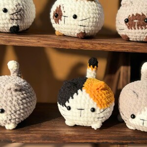 K&ouml;nnte beinhalten: Eine Sammlung handgefertigter H&auml;kelkatzenfiguren in verschiedenen Farben, darunter Wei&szlig;, Grau und Calico-Muster. Jede Katze hat schwarze Knopfaugen und einen kleinen Schwanz. Sie werden auf einem Holzregal ausgestellt.