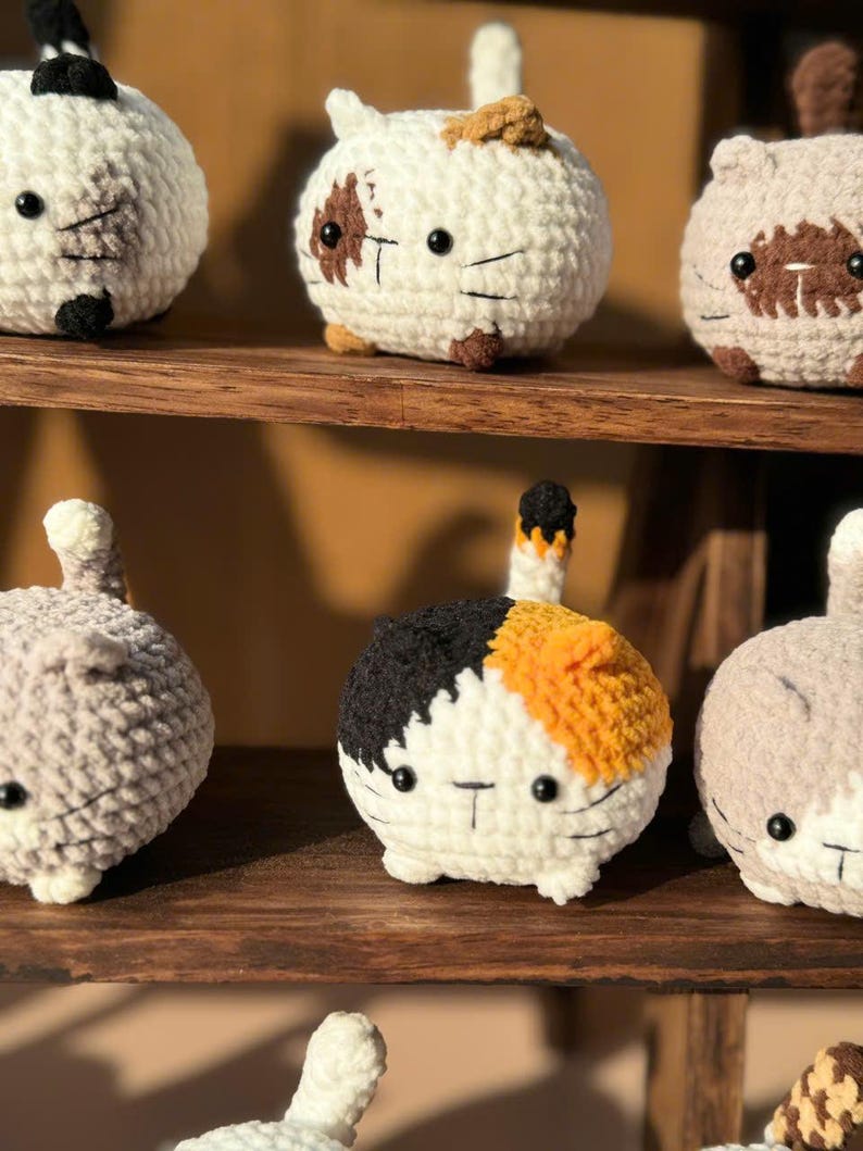 K&ouml;nnte beinhalten: Eine Sammlung handgefertigter H&auml;kelkatzenfiguren in verschiedenen Farben, darunter Wei&szlig;, Grau und Calico-Muster. Jede Katze hat schwarze Knopfaugen und einen kleinen Schwanz. Sie werden auf einem Holzregal ausgestellt.