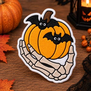 May include: An embroidered patch featuring a bright orange pumpkin with a brown stem, two small black bats with white eyes perched on top, and a beige skeleton hand cradling the pumpkin, all outlined with a thick white border.