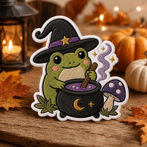 May include: An embroidered patch featuring a green frog wearing a black witch hat with a yellow star, stirring a black cauldron with a crescent moon and star, accompanied by a purple mushroom and swirling purple smoke, with white borders.