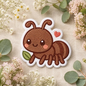 May include: An embroidered patch featuring a brown ant with large eyes, pink cheeks, and a small red heart above its back. The ant is holding a green leaf in its front legs and is outlined in white thread against a neutral fabric background.
