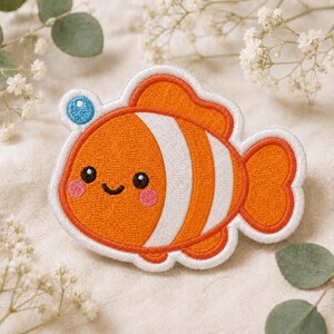 May include: An embroidered patch shaped like a clownfish with bright orange and white stripes. The fish has a small blue bubble above its head, two black eyes, a curved black mouth, and two pink circular cheeks, all outlined in white thread.
