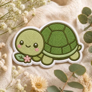 May include: An embroidered patch featuring a light green turtle with a dark green shell, pink cheeks, and a small smile. The turtle is positioned next to a single pink flower with a green leaf, all outlined with a thick white border.