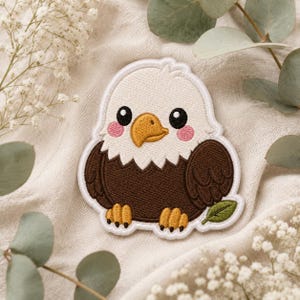 May include: An embroidered patch featuring a cartoon bald eagle with a white head, dark brown body, yellow beak, and yellow feet. The eagle has large black eyes and pink cheeks, with a small green leaf positioned near its right foot.