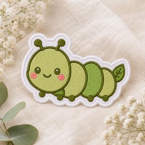May include: An embroidered patch featuring a light green caterpillar with a dark green outline, two antennae, pink cheeks, and a small smile. The segmented body includes a small green leaf detail on the tail and a white border edge.