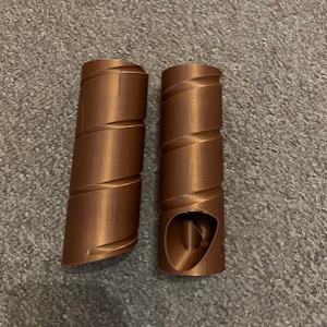 Puede incluir: Dos objetos en espiral de color cobre. Cada uno tiene una forma cilíndrica con una ranura curva continua que recorre su longitud. Un objeto tiene una sección ahuecada en la parte superior, revelando una estructura interna.