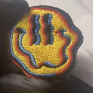 Puede incluir: Parche bordado con una carita sonriente amarilla con un diseño distorsionado. El diseño utiliza hilo rojo, azul y amarillo, creando un efecto trippy y en capas. El parche tiene un borde negro.