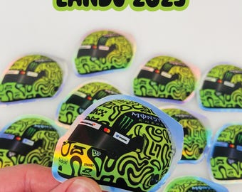 May include: A collection of stickers featuring a racing helmet design with a green and black swirled pattern. The stickers have the text "Lando 2025" in green. The stickers are held by a hand.