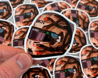 May include: A close-up of a sticker featuring a racing helmet with a snake design. The helmet is white with black and orange accents. The sticker has the text "Google Pixel" and "Tezos" on it.
