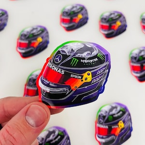 Może przedstawiać: Kolorowa naklejka z kaskiem wyścigowym Formuły 1 z logo Monster Energy, marką Petronas i sponsoringiem Epson. Kask jest w kolorze gradientowym fioletowym, niebieskim, zielonym i czerwonym.