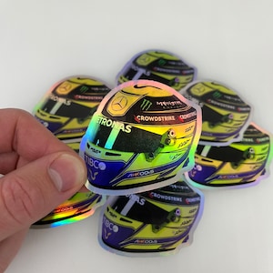 Könnte beinhalten: Ein Set mit sechs holografischen Aufklebern, die einen Formel-1-Helm zeigen. Der Helm ist gelb und schwarz mit einem blauen Visier und verschiedenen Sponsorenlogos, darunter Petronas, Monster Energy, Crowdstrike und Tibco.