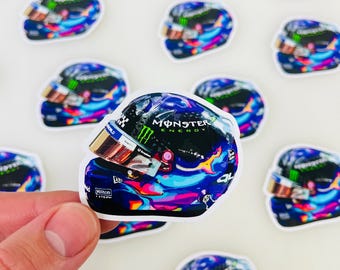 May include: A colorful racing helmet sticker with a Monster Energy logo and a blue, pink, and green paint design. The sticker is on a white background.