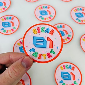 Puede incluir: Una colección de pegatinas redondas con fondo rosa y borde naranja. Las pegatinas presentan el texto "Oscar Pastry" en colores arcoíris y una ilustración azul y blanca de un pastel.