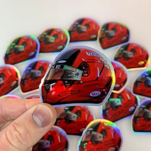 Op de afbeelding: Een verzameling van iriserende stickers met een racecoureur die een rode helm draagt met een wit en blauw ontwerp. De stickers hebben een holografische afwerking en bevatten de handtekening van de coureur, evenals de logo's van verschillende sponsors, waaronder "DBWT", "Castrol", "Amazon Music" en "Bell".