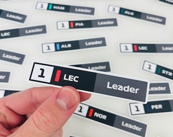 Könnte beinhalten: Nahaufnahme einer Hand, die einen schwarzen und grauen Aufkleber mit der Nummer 1, den Buchstaben LEC und dem Wort Leader hält. Der Aufkleber ist einer von vielen ähnlichen Aufklebern mit verschiedenen Fahrernamen und Nummern.