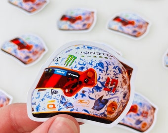 Puede incluir: Un casco de carreras blanco con un patrón floral azul y blanco, una visera roja y un logotipo de Monster Energy. El casco tiene un logotipo de New Era en el lateral.