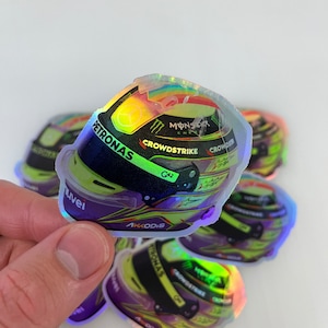Może przedstawiać: Holograficzna naklejka przedstawiająca zielono-fioletowy kask wyścigowy z napisami "Petronas", "Monster Energy", "Crowdstrike", "G42" i "Akodis".