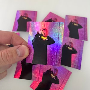 Può includere: Un set di otto adesivi olografici con una persona che indossa un casco rosa e blu con un bagliore giallo. Gli adesivi sono quadrati e hanno uno sfondo rosa e blu con un effetto glitter.