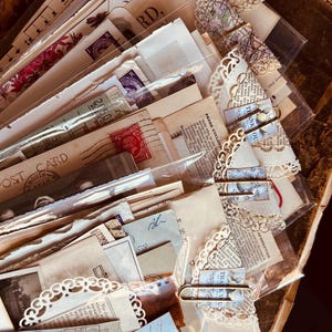 May include: A selection of vintage ephemera, including postcards, stamps, and paper scraps, arranged in clear plastic sleeves. Some items are secured with decorative paper clips. The papers have a variety of colours and textures, with visible text and floral designs.
