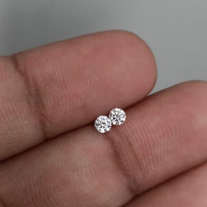 May include: Two small, round, brilliant-cut diamonds. The diamonds are clear and sparkling, with multiple facets reflecting light. The gems are displayed on a skin-toned background.
