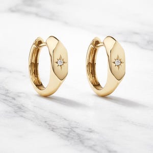 Könnte beinhalten: Ein Paar goldene Creolen. Jeder Ohrring hat ein geometrisches Design mit einem kleinen sternförmigen Diamantakzent. Die Ohrringe werden auf einer weiß-grauen Marmoroberfläche präsentiert.