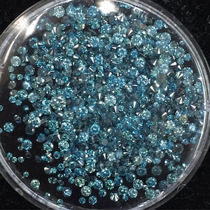 May include: A close-up view of a small plastic container filled with loose blue diamonds. The diamonds are various sizes and shapes.