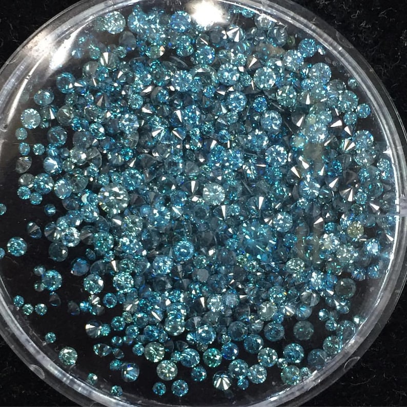 May include: A close-up view of a small plastic container filled with loose blue diamonds. The diamonds are various sizes and shapes.