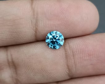 May include: A round, faceted, brilliant-cut gemstone in a vibrant shade of light blue. The gem is held in a hand, showcasing its clarity and sparkle. The gemstone's facets reflect light, creating a dazzling display of color and brilliance.