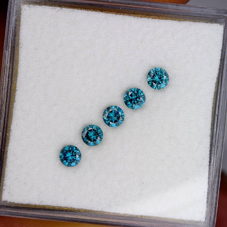 May include: Five round blue gemstones are displayed in a row on a white surface inside a small box. The gemstones are arranged in a diagonal line from top right to bottom left.