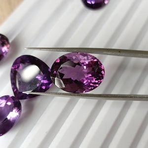May include: A close-up of a purple amethyst gemstone held by tweezers. The gemstone is oval-shaped and has a faceted cut. The gemstone is surrounded by other amethyst gemstones on a white surface.