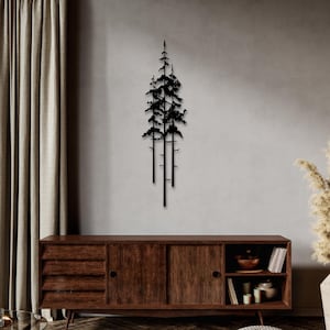 Op de afbeelding: Zwarte metalen wandkunst met een silhouet van drie dennenbomen. De kunst is gemonteerd op een grijze muur boven een houten dressoir.