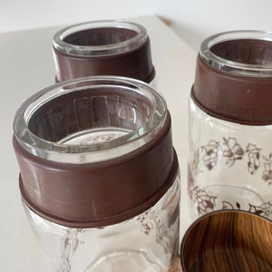 May include: Three glass jars with brown lids and floral designs. The jars are clear glass and have a brown rim around the top. The lids are a dark brown color and have a textured surface.