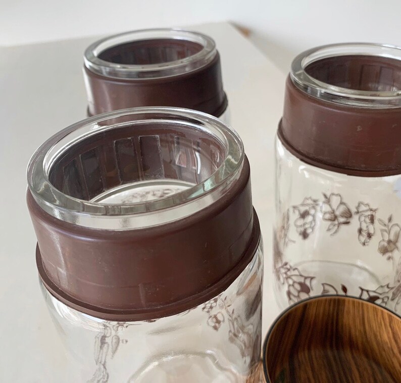 May include: Three glass jars with brown lids and floral designs. The jars are clear glass and have a brown rim around the top. The lids are a dark brown color and have a textured surface.