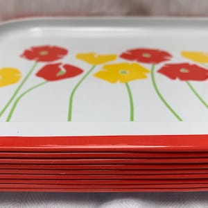 Op de afbeelding: Een stapel rode en witte metalen dienbladen met een bloemenmotief. De dienbladen hebben gele en rode bloemen met groene stelen.