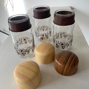 May include: Three clear glass jars with brown lids and a grapevine design. The jars have wooden lids with a woodgrain pattern.