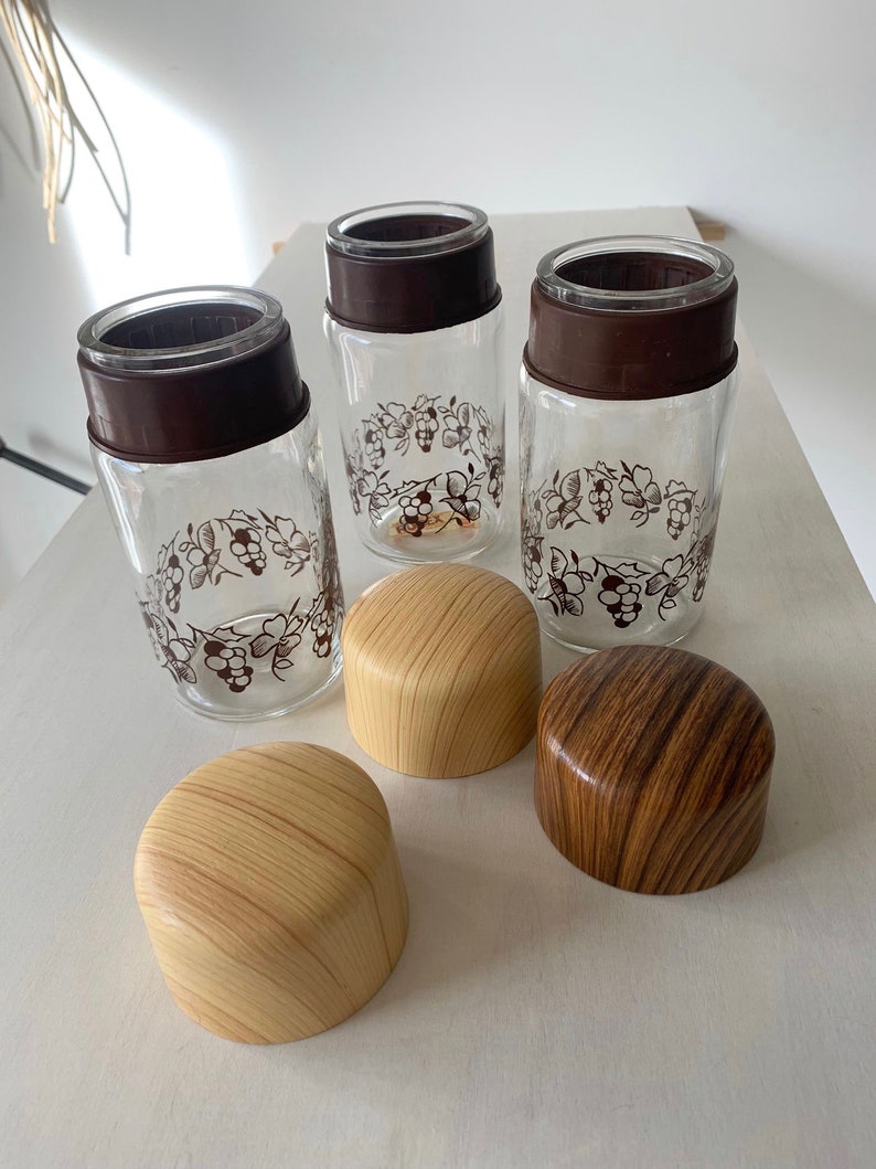 May include: Three clear glass jars with brown lids and a grapevine design. The jars have wooden lids with a woodgrain pattern.