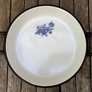 Peut inclure: Poêle en fonte émaillée blanche avec poignées et bord noirs. Un motif floral bleu est centré à l'intérieur. La poêle a deux grandes poignées noires en forme de D.