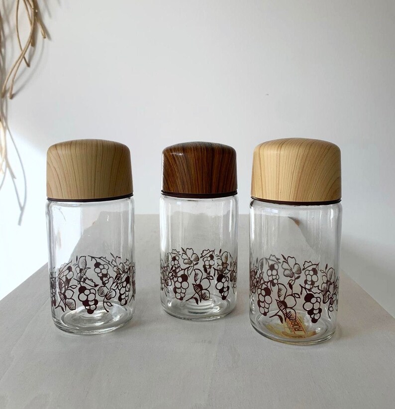 May include: Three glass jars with wooden lids and a grapevine design. The jars are clear glass and the lids are a light brown wood.