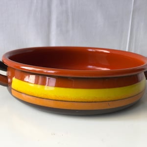 Puede incluir: Un bol de servir de cerámica marrón con dos asas. El bol tiene un diseño a rayas en naranja, amarillo y marrón.