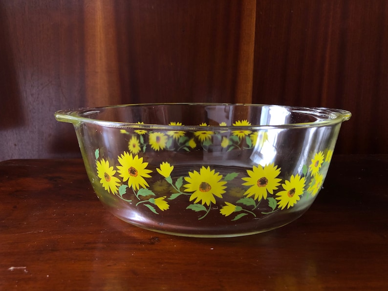 May include: A clear glass baking dish with a yellow and green floral design. The design features a repeating pattern of sunflowers with green leaves.