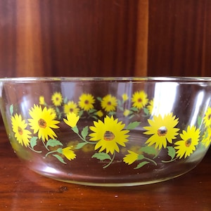 May include: A clear glass baking dish with a yellow and green sunflower design. The dish has a handle on one side.