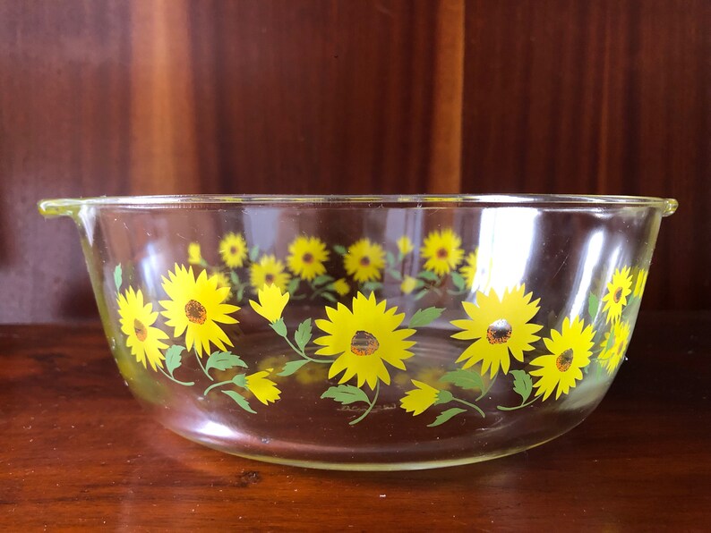May include: A clear glass baking dish with a yellow and green sunflower design. The dish has a handle on one side.