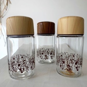 May include: Three glass jars with wooden lids and a grapevine design. The jars are clear glass with a brown grapevine design around the middle. The lids are a light wood color.